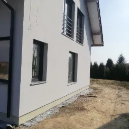 Świeżo otynkowana, jasnoszara elewacja domu jednorodzinnego z grafitowymi oknami i metalowymi balustradami, widok z boku na tle nieba i drzew.