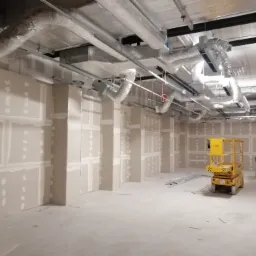 Wnętrze w trakcie prac wykończeniowych: ściany obłożone płytami gipsowo-kartonowymi, widoczne łączenia szpachlowane, instalacja wentylacyjna pod sufitem, podnośnik nożycowy w tle.