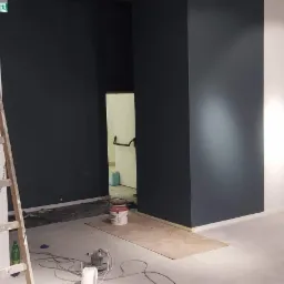 Wnętrze w trakcie malowania: ściany pomalowane w odcieniach szarości i bieli, drewniana drabina oparta o ścianę, puszki z farbą na podłodze, widoczne kable i akcesoria malarskie.