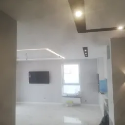 Nowoczesne wnętrze z białymi marmurowymi płytkami podłogowymi, oświetleniem sufitowym LED i czarnym telewizorem na ścianie, widok na okno z grzejnikiem i akcesoriami.