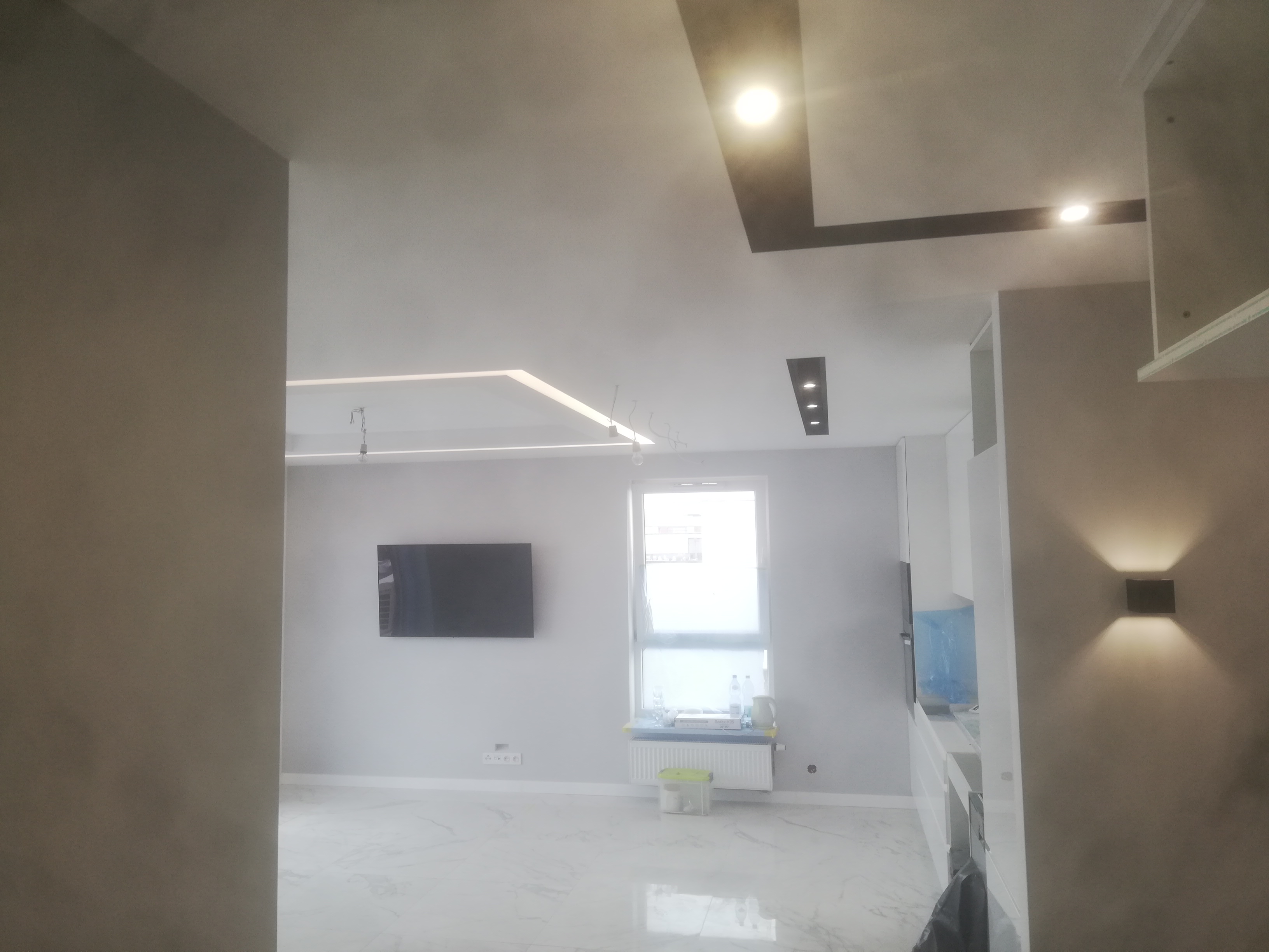 Nowoczesne wnętrze z białymi marmurowymi płytkami podłogowymi, oświetleniem sufitowym LED i czarnym telewizorem na ścianie, widok na okno z grzejnikiem i akcesoriami.