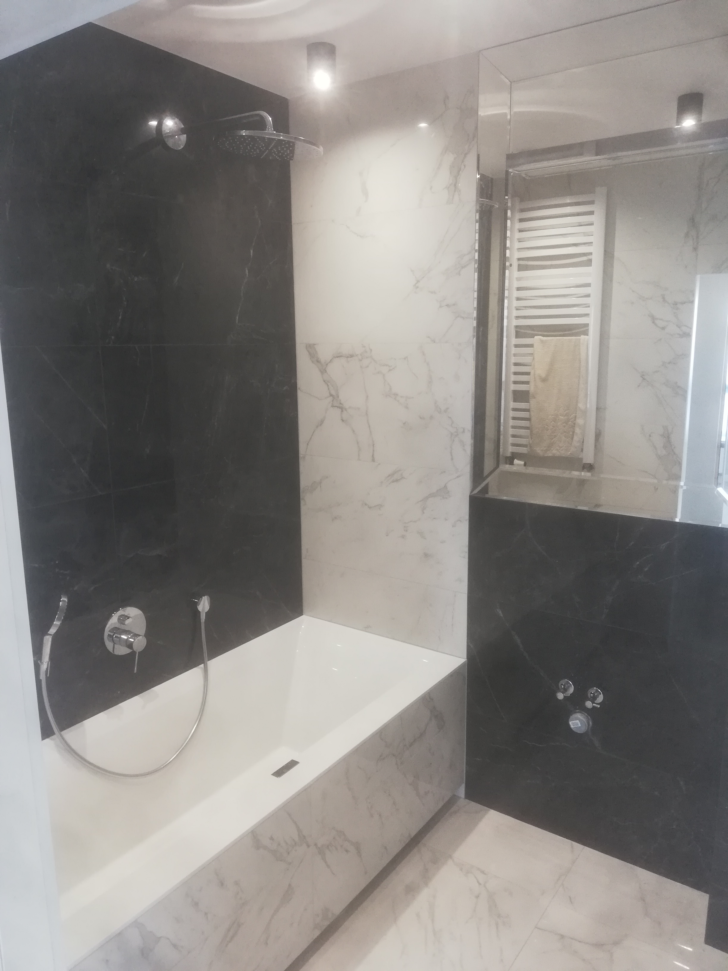 Nowoczesna łazienka z czarno-białymi płytkami imitującymi marmur, deszczownicą i grzejnikiem drabinkowym widocznym w lustrze.