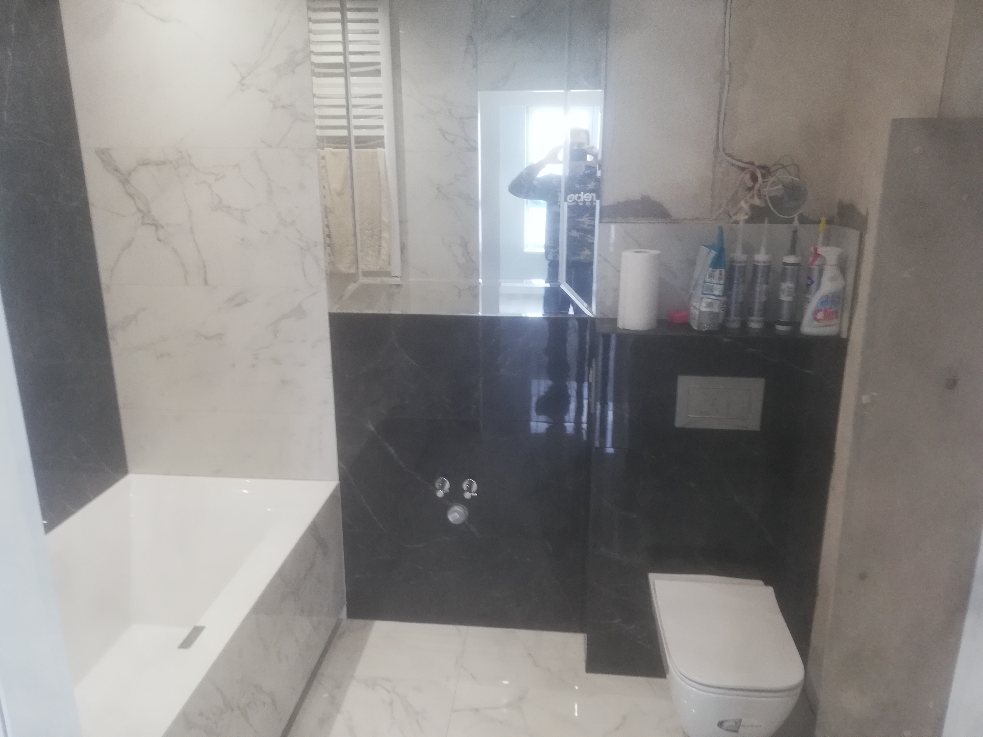 Łazienka w trakcie wykańczania: biała wanna i toaleta, czarne płytki imitujące marmur na ścianach, widoczne rury i instalacje, odbicie fotografa w lustrze.