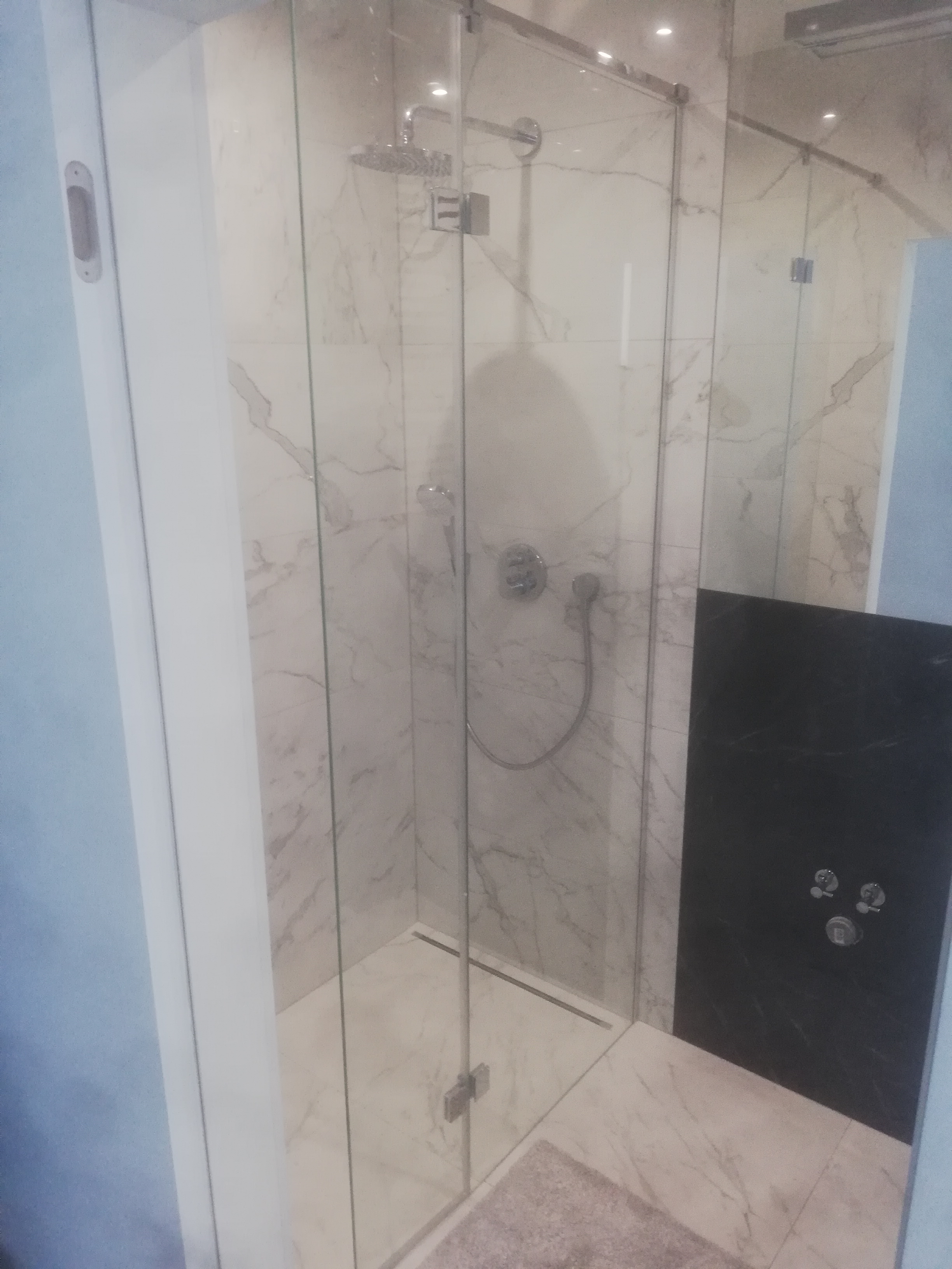 Kabina prysznicowa z deszczownicą i baterią termostatyczną, wykończona płytkami imitującymi marmur Carrara, z czarnym akcentem na ścianie, widok z boku.