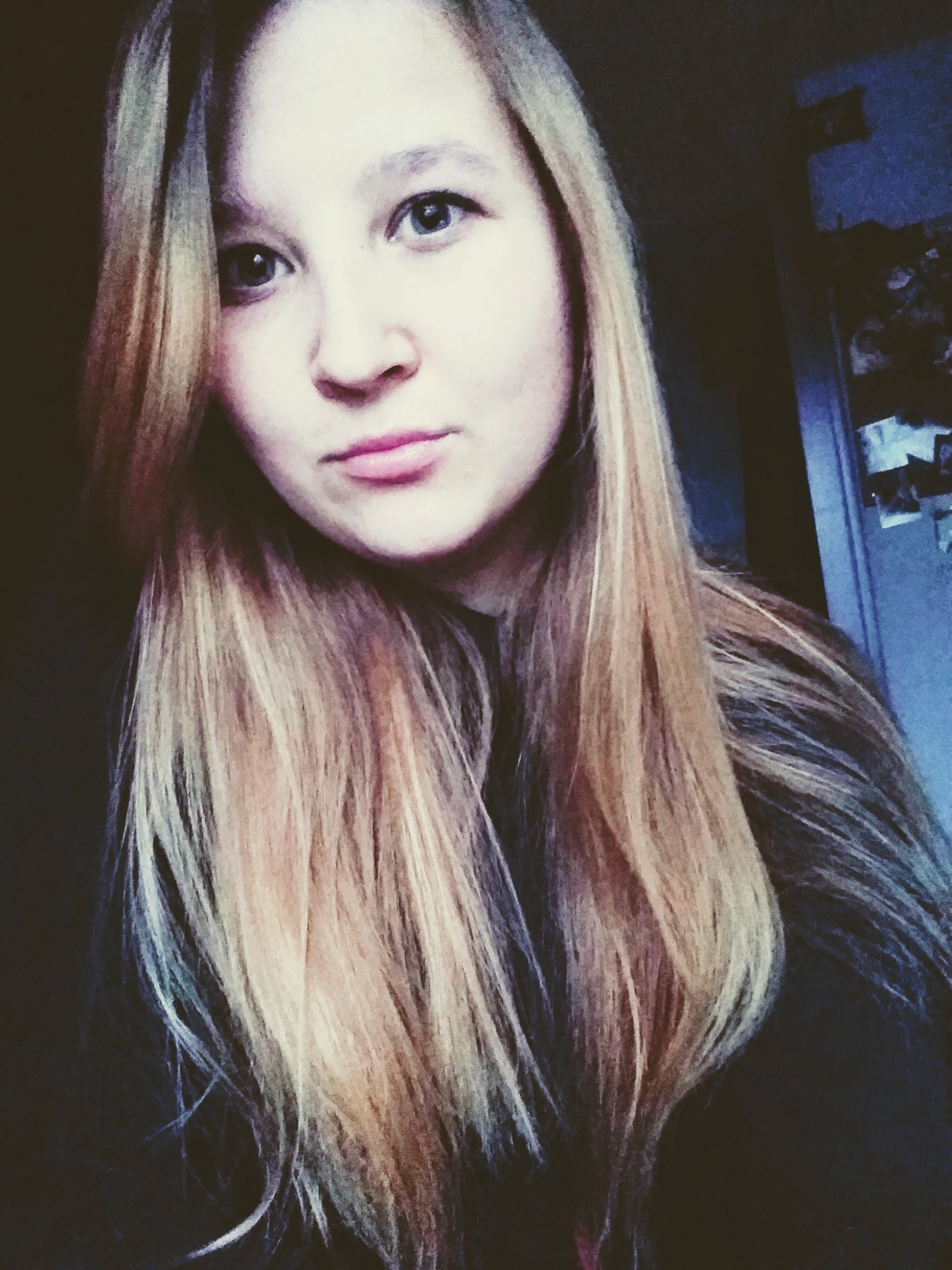 Portret młodej kobiety z długimi, blond włosami, z delikatnym uśmiechem i niebieskimi oczami, sfotografowana w słabym oświetleniu z widocznymi zdjęciami przypiętymi do ściany w tle.