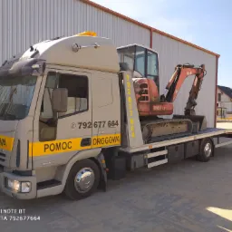 Laweta z żółtymi napisami 'POMOC DROGOWA' i numerem telefonu przewożąca małą, czerwoną koparkę O&K na tle magazynu i budynku w budowie.