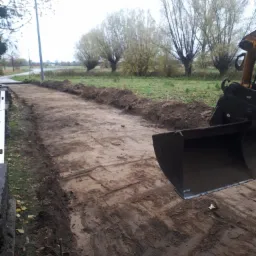 Przygotowanie terenu pod drogę: wyrównany pas ziemi z widocznymi śladami opon i łyżką koparki w kadrze, w tle rzadkie drzewa i pochmurne niebo.