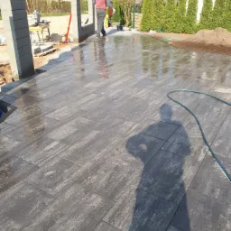 Układanie mokrej, prostokątnej kostki brukowej w odcieniach szarości, widoczny pracownik z przecinarką do kostki i słupki ogrodzeniowe w tle.