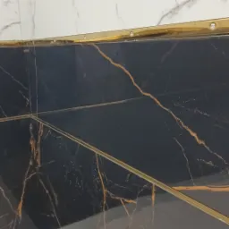 Częściowo ułożone płytki: białe marmurowe na górze i czarne z brązowymi żyłami na dole, oddzielone złotą listwą, widoczne wystające przewody elektryczne.