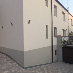 MURTOR Piotr Opiński - Dwukondygnacyjny budynek z tarasem nad garażem, otynkowany na biało z szarym pasem u dołu, z kamienną kostką brukową na podjeździe i oświetleniem zewnętrznym.