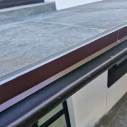 Szare płytki na tarasie lub balkonie, detale wykończenia krawędzi z brązowym profilem i widoczną uszczelką, ujęcie z bliska.