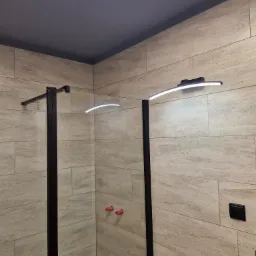 Kabina prysznicowa z czarną ramą i oświetleniem LED, zamontowana w łazience wyłożonej beżowymi, podłużnymi płytkami imitującymi kamień, widoczne wystające czerwone rurki instalacyjne w ścianie.