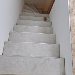 MURTOR Piotr Opiński - Wykończone schody wewnętrzne z jasnymi, kamiennymi płytkami i srebrnymi listwami krawędziowymi, widok z góry. Na jednym ze stopni leży cegła.