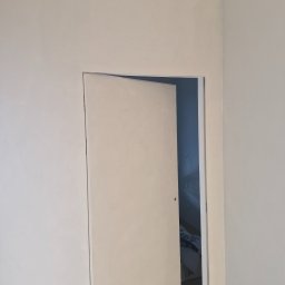 MURTOR Piotr Opiński - Otwarta, beżowa, minimalistyczna ściana z ukrytymi drzwiami w pokoju. Widoczny fragment wnętrza z pościelą w tle. Świeżo pomalowane ściany w neutralnych barwach.