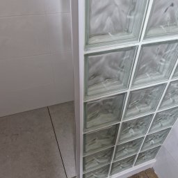 MURTOR Piotr Opiński - Szklana ścianka działowa z luksferów w łazience, widok z bliska na połączenie z podłogą i ścianą. Minimalistyczny design, jasne płytki.