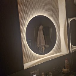 MURTOR Piotr Opiński - Wykończona łazienka w Toruniu: ściana z dekoracyjną glazurą, okrągłe lustro z podświetleniem LED, nowoczesna umywalka z armaturą i akcesoriami.