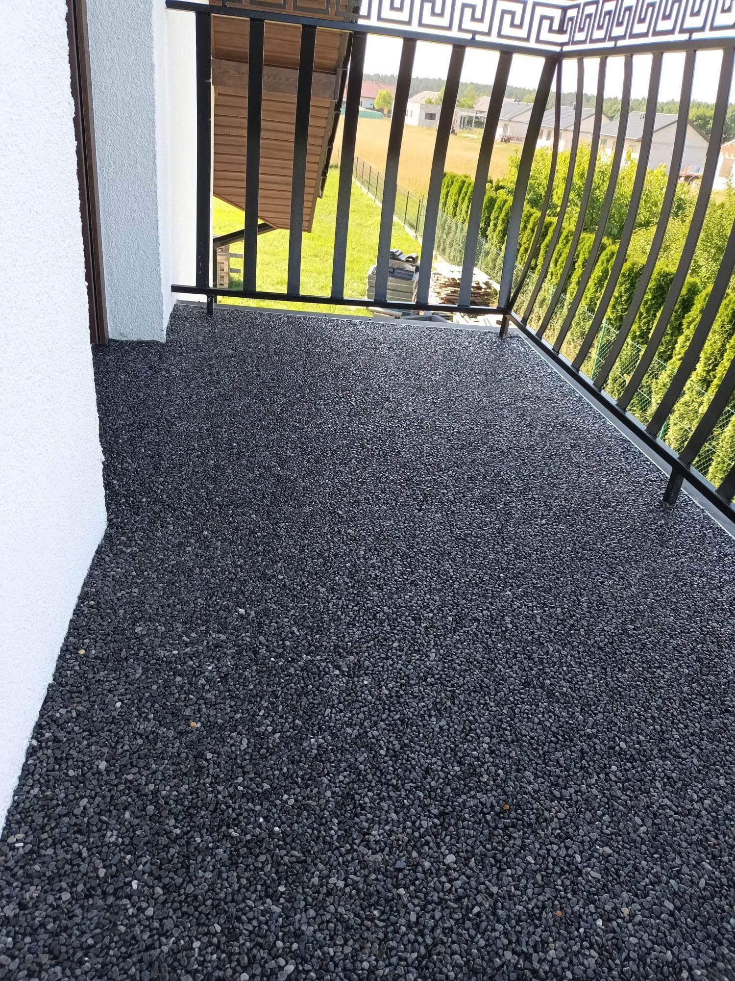 Czarna, drobna posadzka kamienna na balkonie z metalową balustradą o geometrycznym wzorze, widok na zielony ogród i zabudowania w tle.