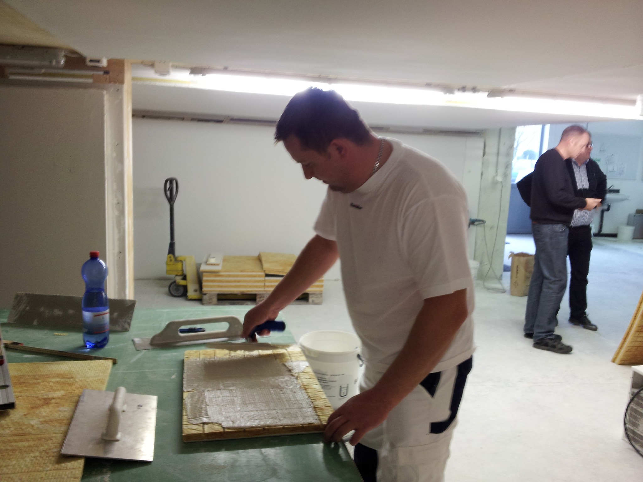 Pracownik rozprowadza klej na płytce za pomocą szpachelki, przygotowując ją do montażu, w tle widoczni dwaj mężczyźni i paleta z płytkami.