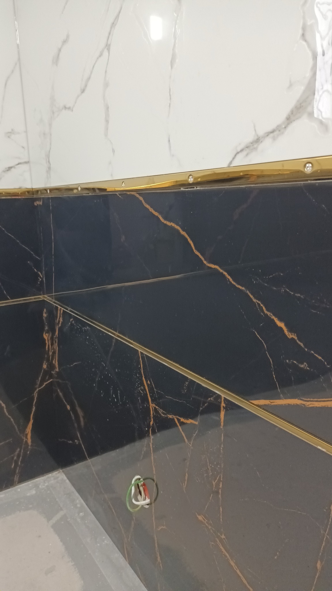 Częściowo ułożone płytki: białe marmurowe na górze i czarne z brązowymi żyłami na dole, oddzielone złotą listwą, widoczne wystające przewody elektryczne.
