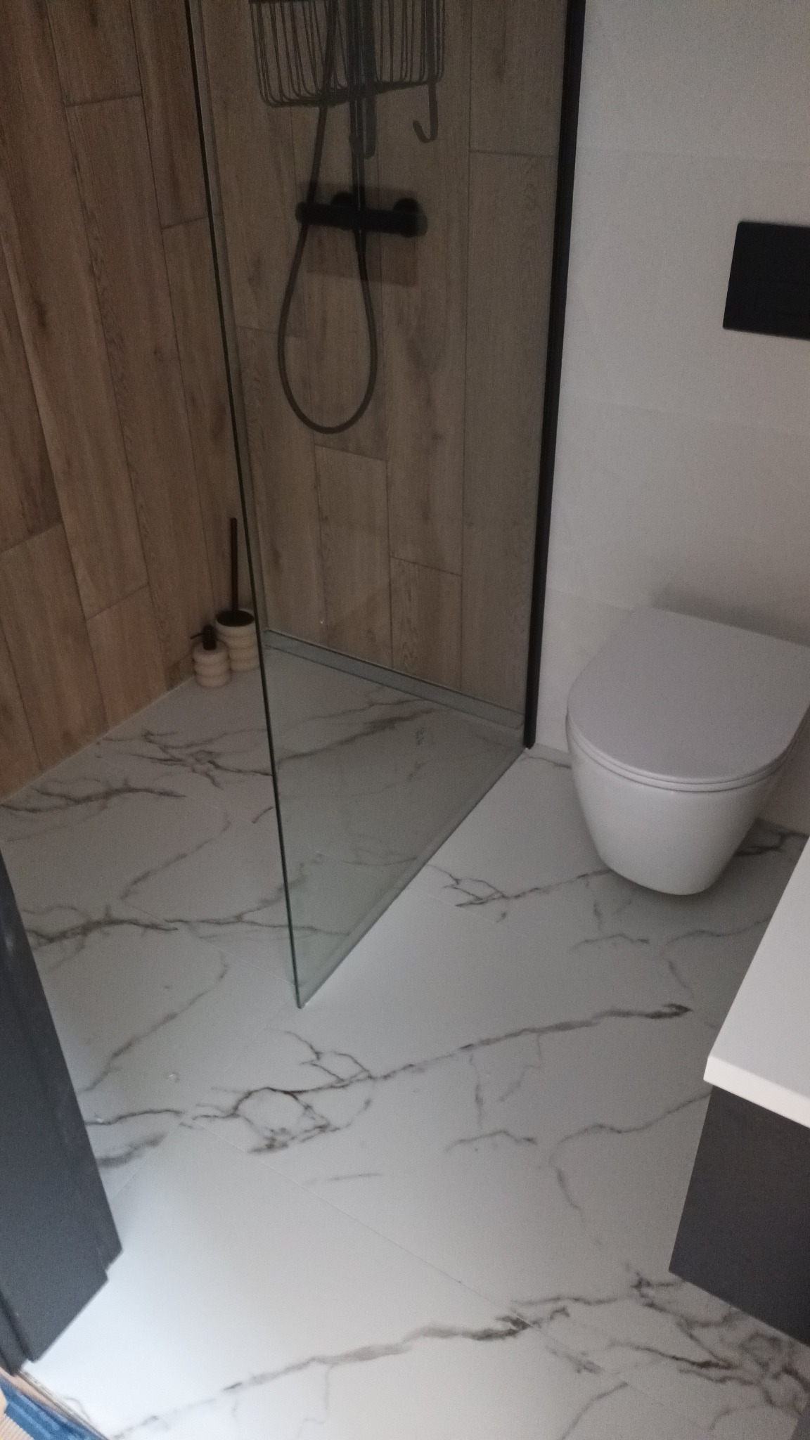 Nowoczesna łazienka z kabiną prysznicową typu walk-in, białą armaturą i płytkami imitującymi marmur na podłodze i ścianach kabiny, widoczny czarny zestaw prysznicowy.