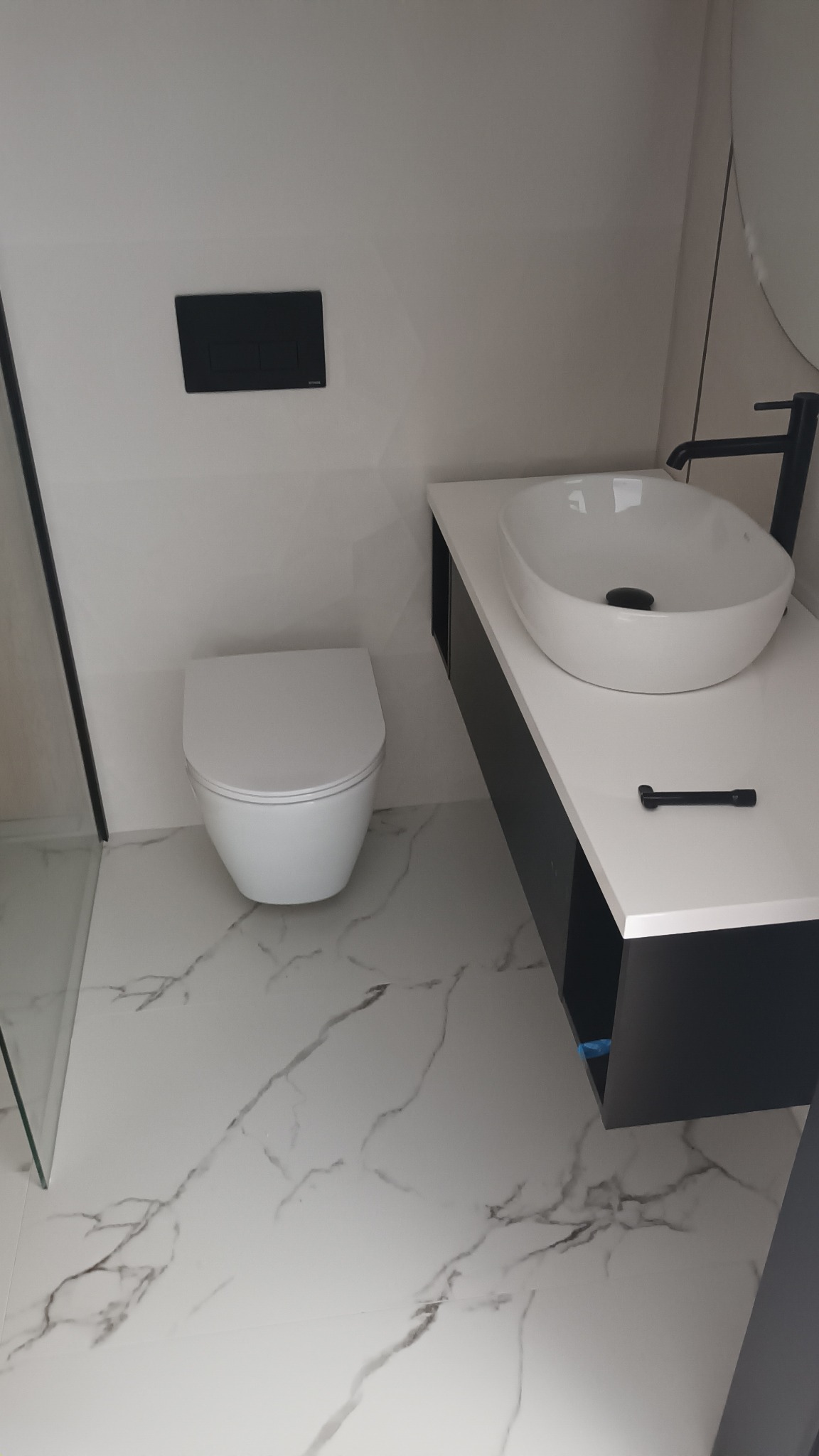 Nowoczesna łazienka z białą umywalką nablatową, czarną armaturą i wiszącą toaletą na tle marmurowych płytek podłogowych.