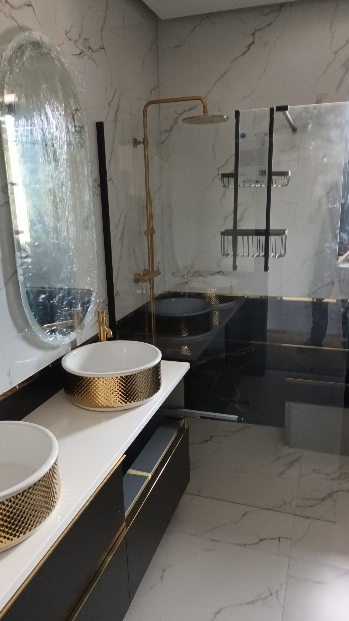 Nowoczesna łazienka z marmurowymi płytkami, złotą armaturą i umywalkami nablatowymi z dekoracyjnym, złotym wzorem, lustro w folii ochronnej przed montażem.