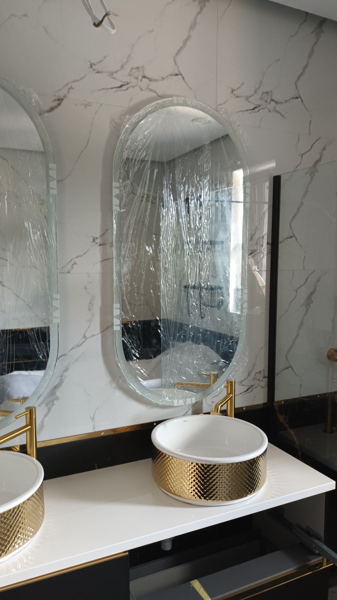 Dwie owalne umywalki nablatowe z białym wnętrzem i złotą, teksturowaną obudową, zamontowane na białym blacie z czarnymi akcentami, pod lustrami zabezpieczonymi folią ochronną na tle marmurowej ściany.