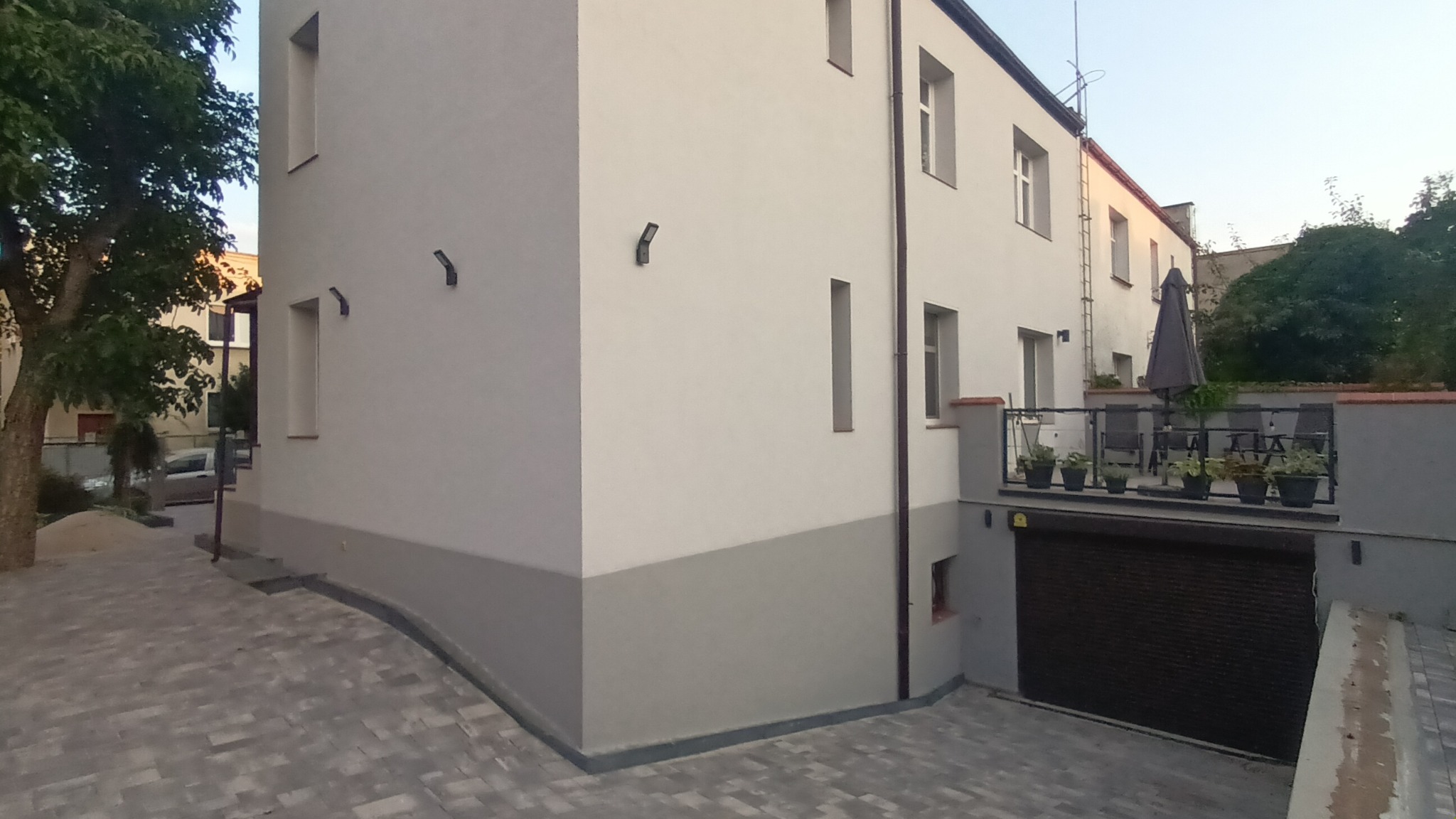 Dwukondygnacyjny budynek z tarasem nad garażem, otynkowany na biało z szarym pasem u dołu, z kamienną kostką brukową na podjeździe i oświetleniem zewnętrznym.