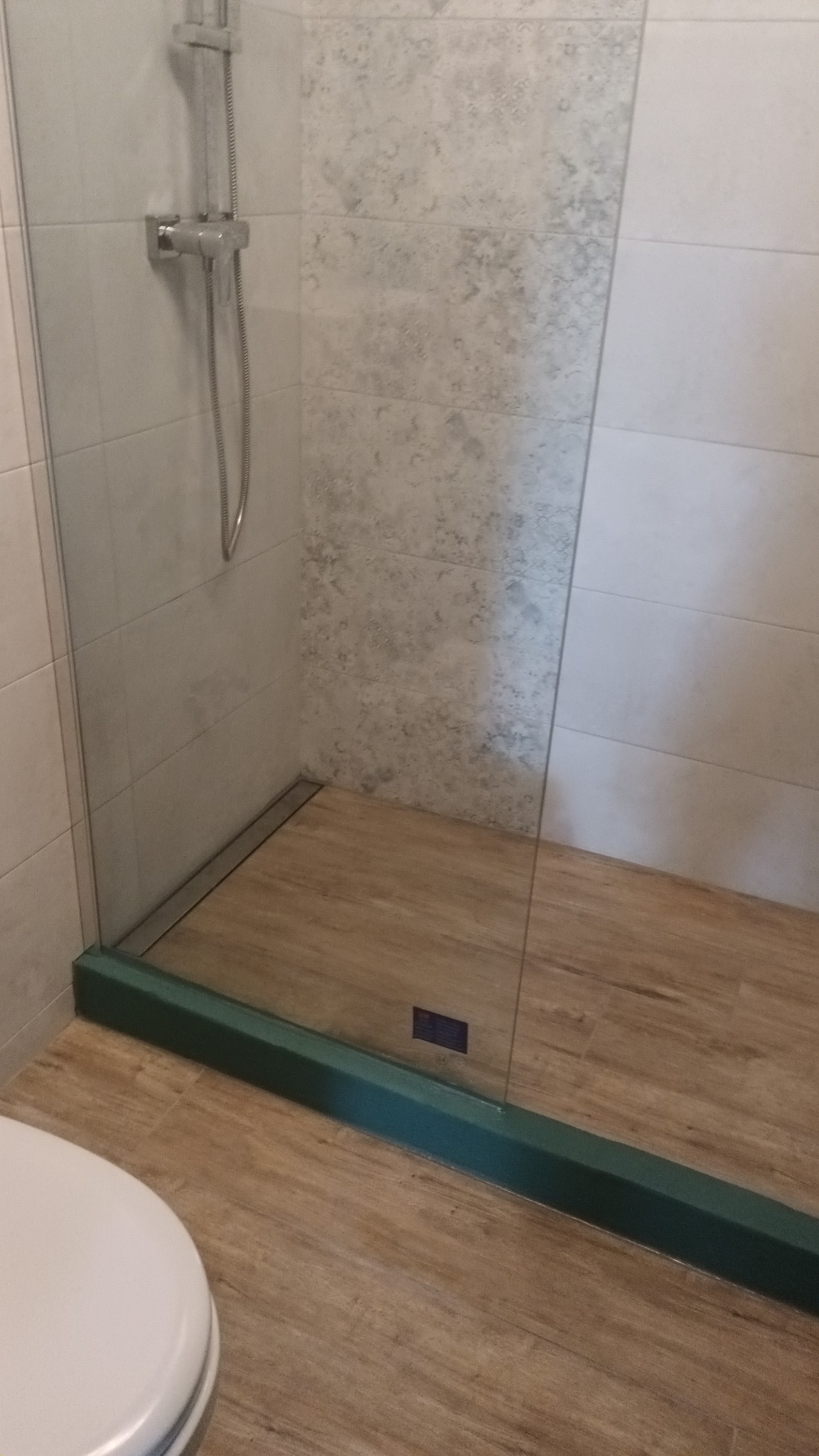 Nowoczesna kabina prysznicowa walk-in z odpływem liniowym, wykończona płytkami imitującymi drewno na podłodze i szaro-białymi płytkami na ścianach, z widocznym fragmentem toalety w dolnym lewym rogu.