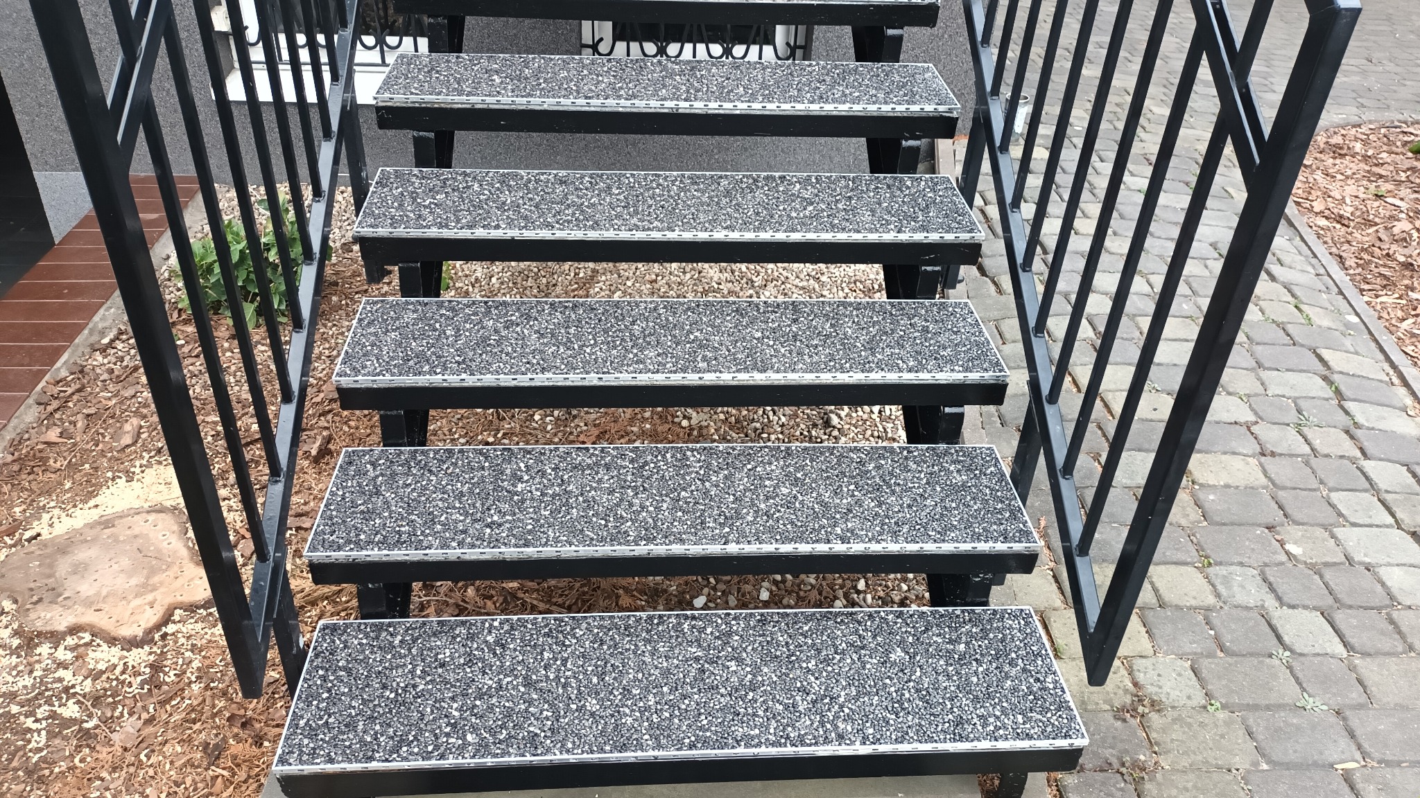Zewnętrzne schody z czarną metalową balustradą i powierzchnią stopni wykończoną szarą, antypoślizgową posypką z kamyczków, widok z dołu na tle kostki brukowej i kory.