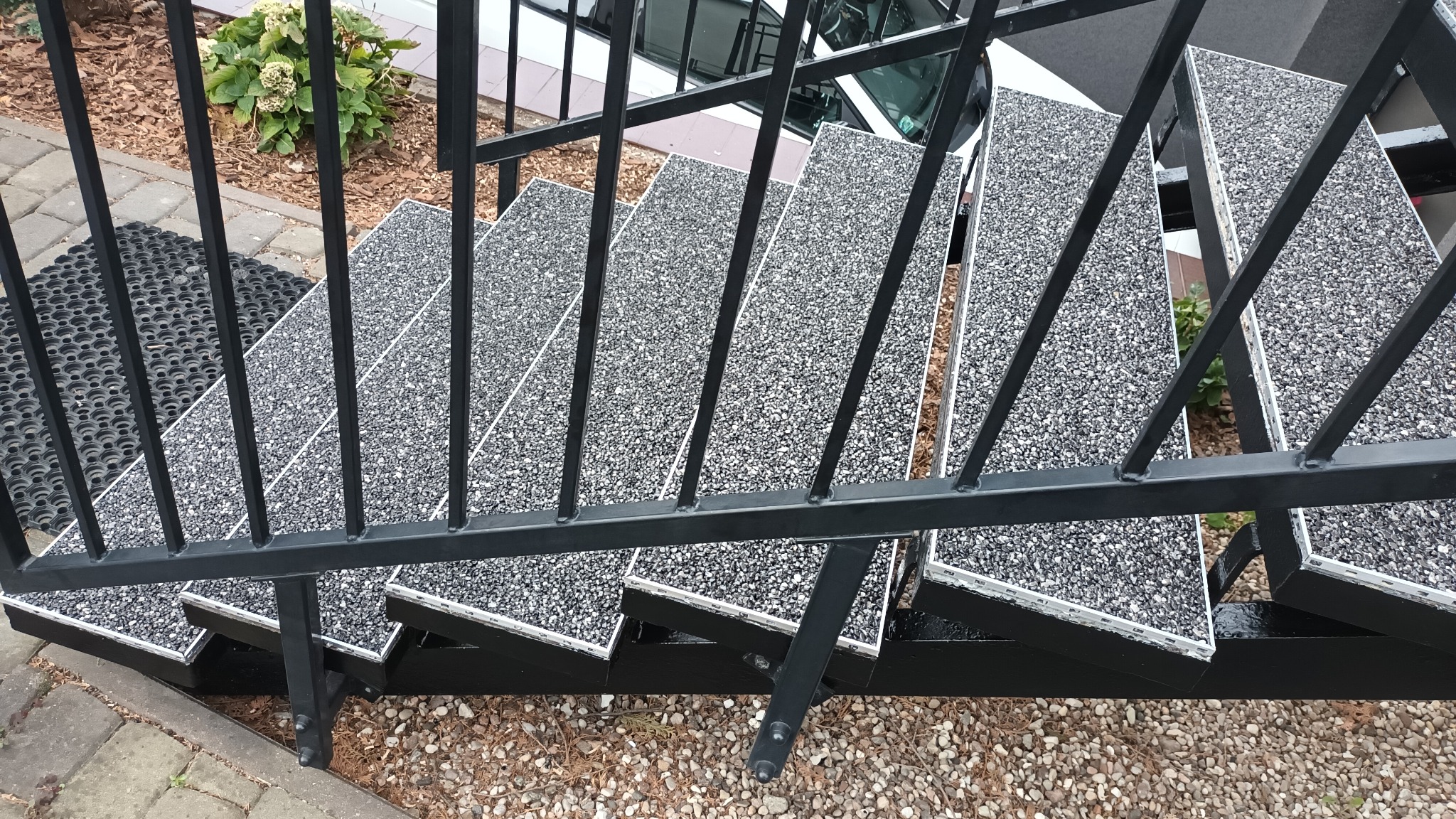Schody zewnętrzne z czarną metalową balustradą i powierzchnią stopni wykończoną szarą, drobną mieszanką kamieni, widoczne z góry pod kątem, obok krzew i mata gumowa