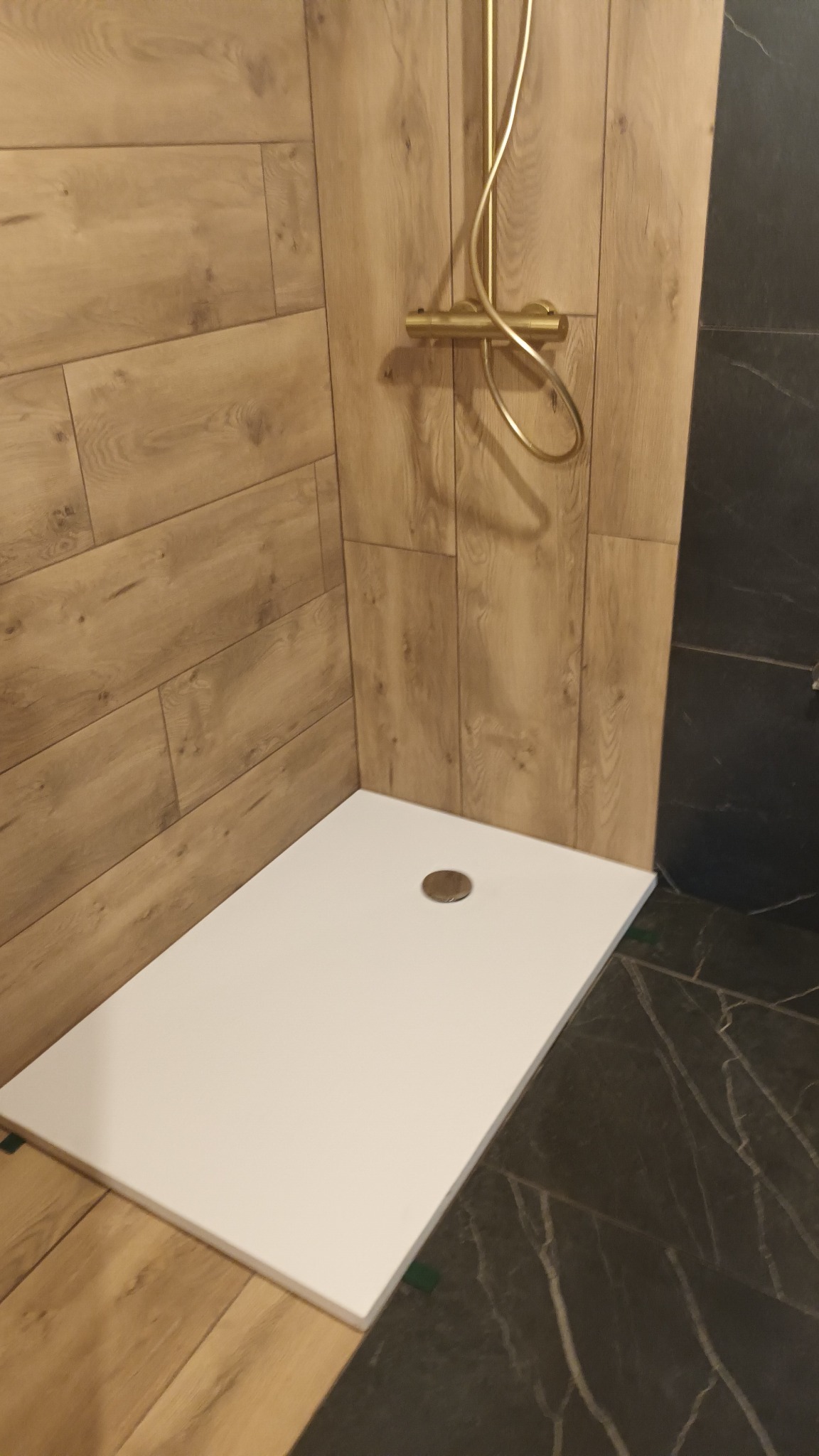Nowoczesny brodzik z odpływem w rogu łazienki, otoczony panelami ściennymi imitującymi drewno oraz płytkami w odcieniach szarości z marmurkowym wzorem, z widoczną złotą baterią prysznicową.