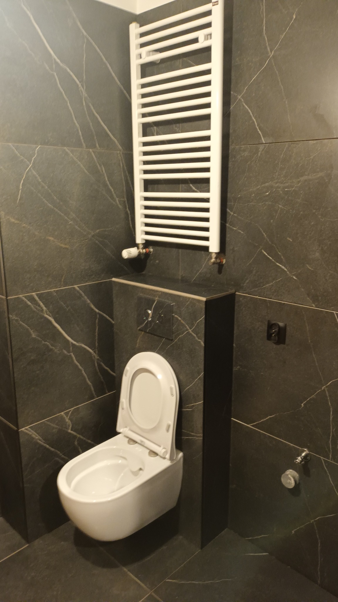 Nowoczesna łazienka z ciemnymi płytkami imitującymi marmur, wisząca toaleta z otwartą klapą i biały grzejnik drabinkowy na ścianie.
