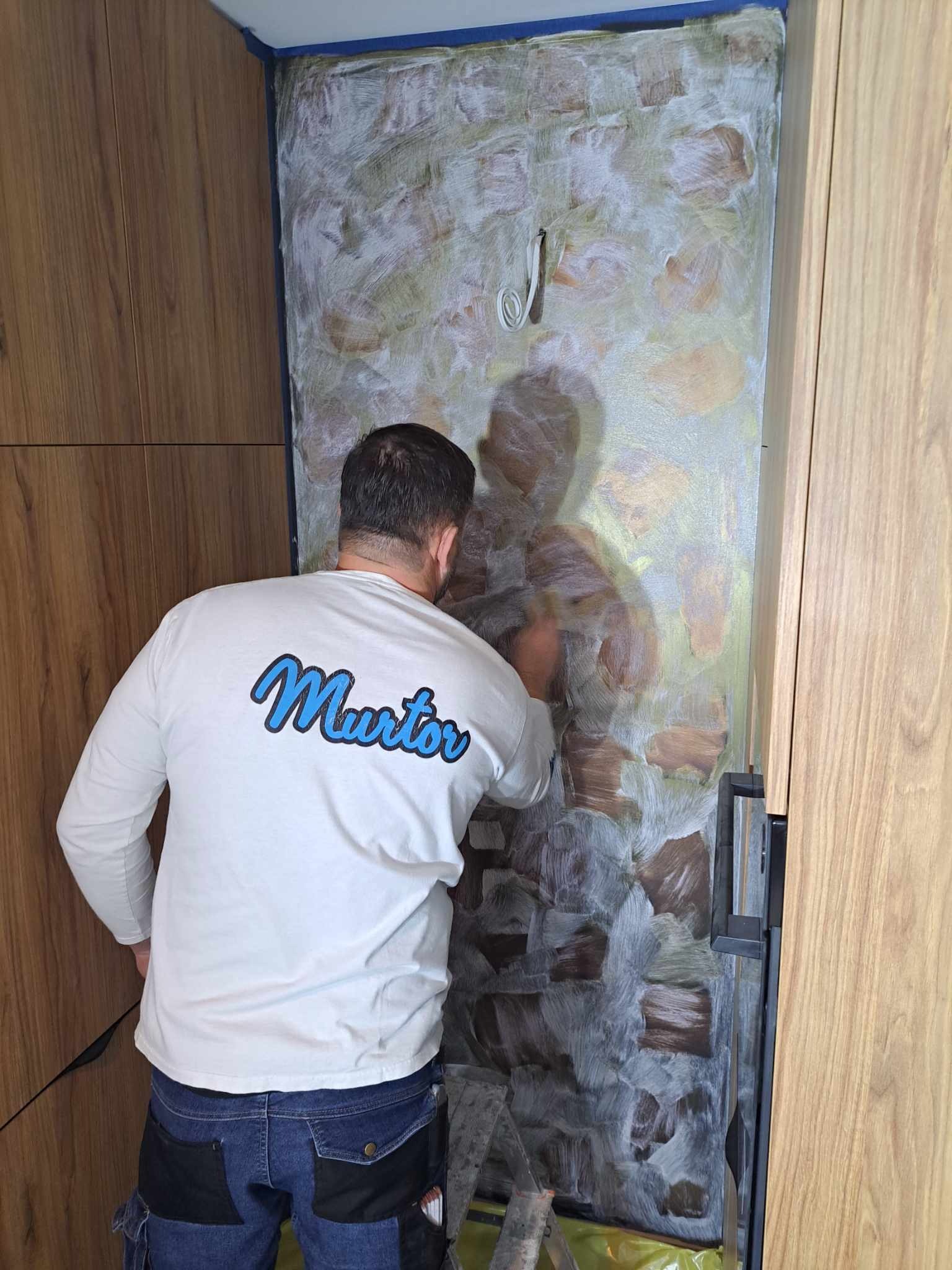 Mężczyzna w koszulce z logo firmy nakłada szpachelką dekoracyjną masę na ścianę w łazience, zabezpieczoną niebieską taśmą malarską, widoczne drewniane elementy zabudowy.