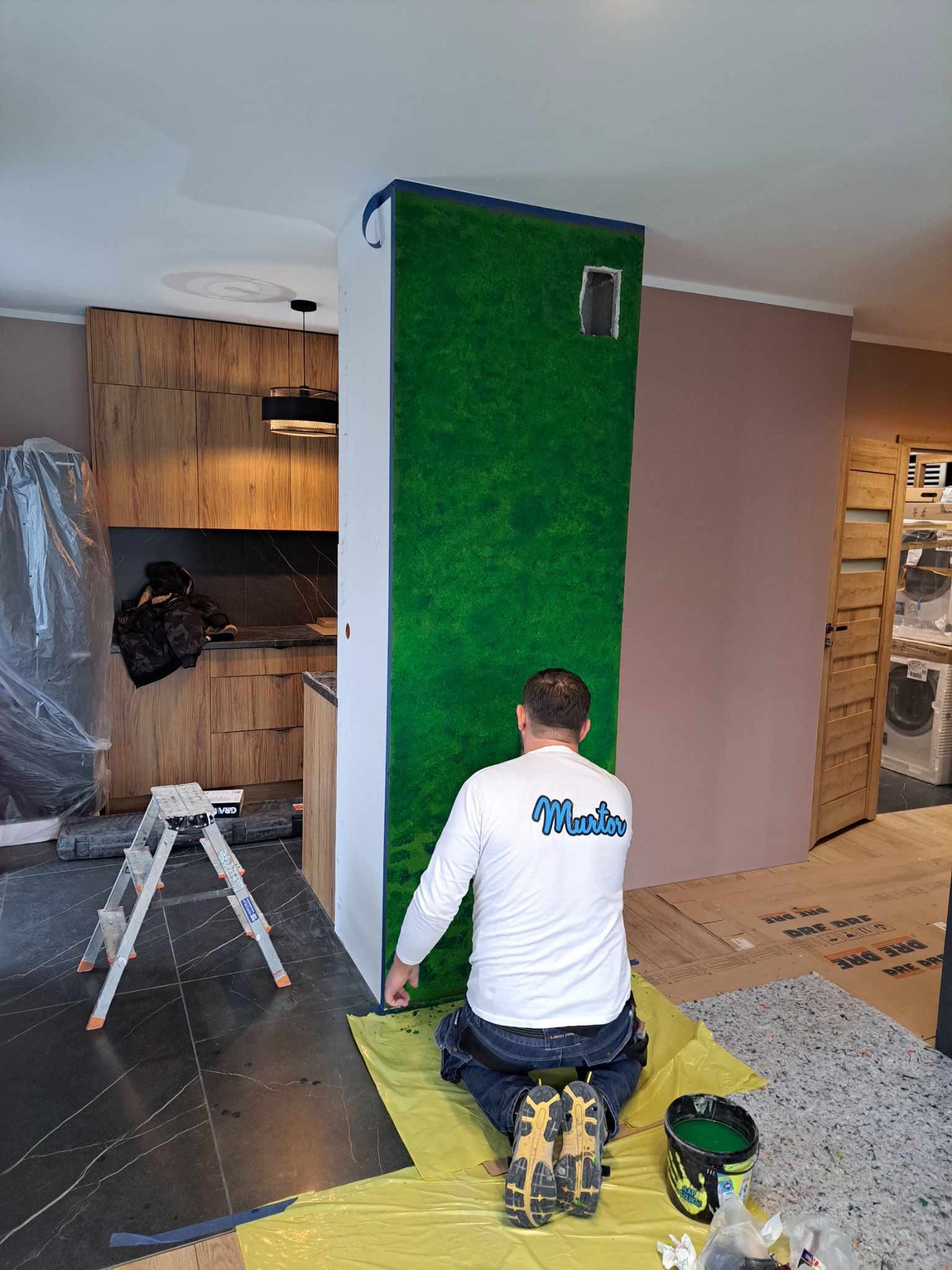 Pracownik na kolanach maluje na zielono fragment ściany z otworem, zabezpieczony niebieską taśmą malarską, widoczna drabina, podłoga wyłożona ciemnymi płytkami imitującymi marmur, kuchnia...