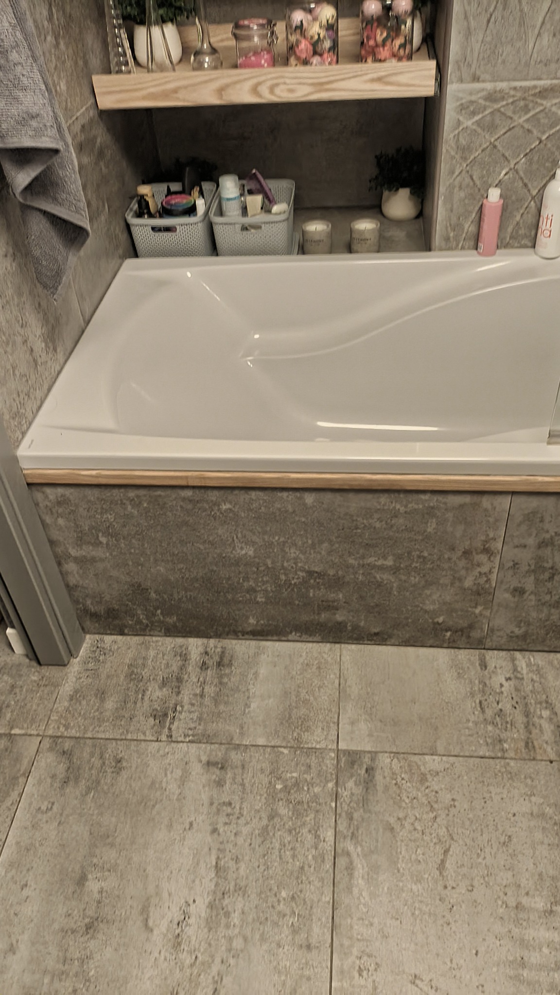 Nowoczesna wanna obudowana szarymi płytkami imitującymi beton, z drewnianym wykończeniem krawędzi, na tle płytek podłogowych w podobnym odcieniu, z półką nad wanną z dekoracjami i kosmetykami.