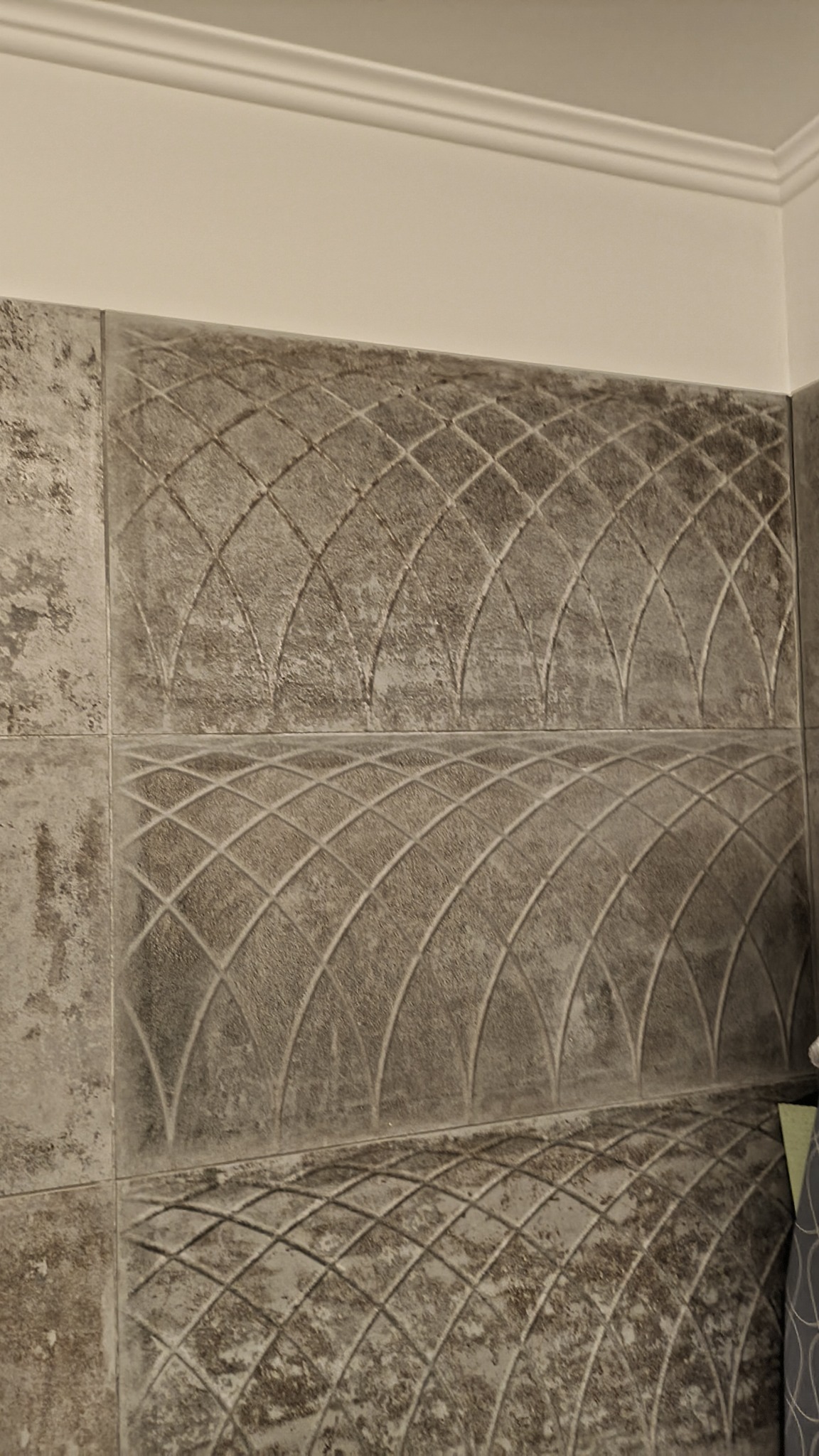 Ściana wyłożona szarymi płytkami z wytłoczonym wzorem łuku i siatki, wykończona białym, ozdobnym gzymsem sufitowym.