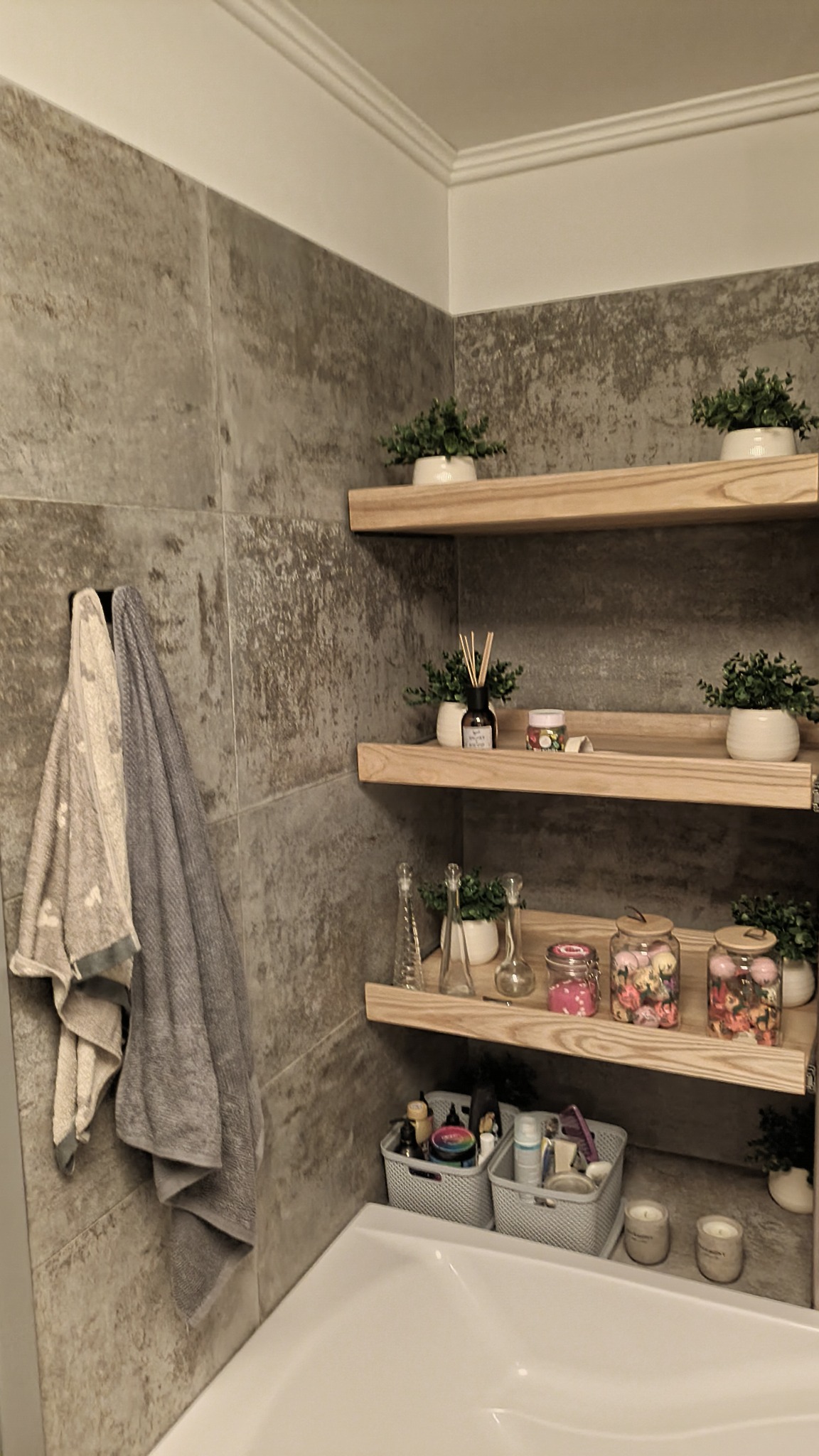Jasnoszara łazienka z półkami z jasnego drewna, na których stoją rośliny w doniczkach, dekoracyjne słoiki i dyfuzor zapachowy, ręczniki wiszące na wieszaku na ścianie z szarych płytek, poniżej...