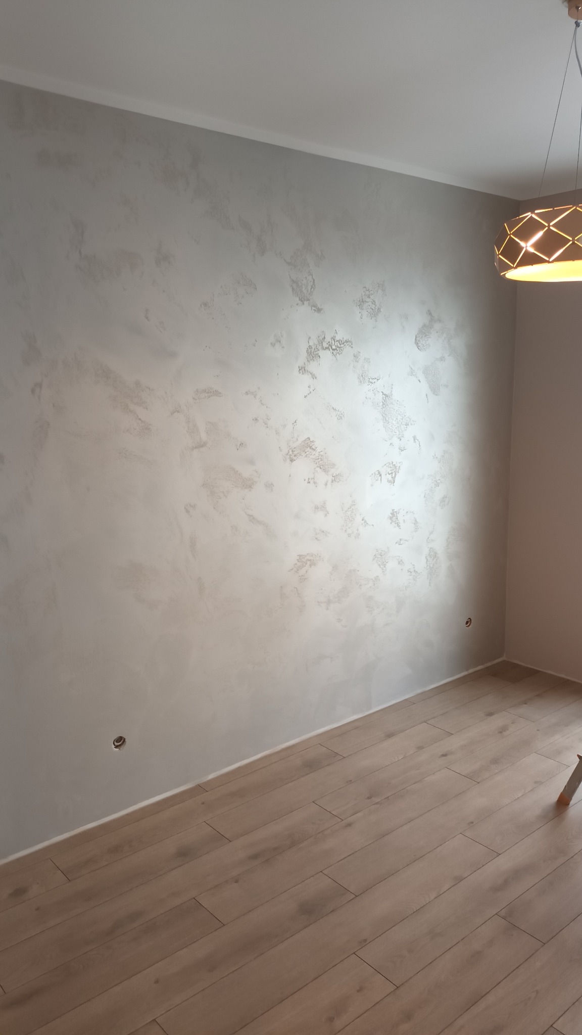 Jasnoszara ściana z dekoracyjną fakturą imitującą chmurki, drewniana podłoga i nowoczesna lampa wisząca z ażurowym kloszem w pustym pokoju.