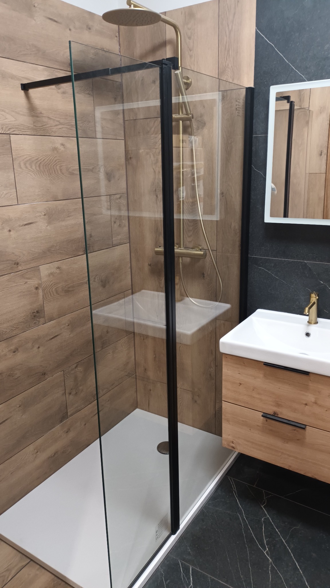 Nowoczesna kabina prysznicowa z czarnymi ramami i złotą armaturą w łazience w Toruniu, z drewnopodobnymi płytkami na ścianach i ciemnym marmurem na podłodze.