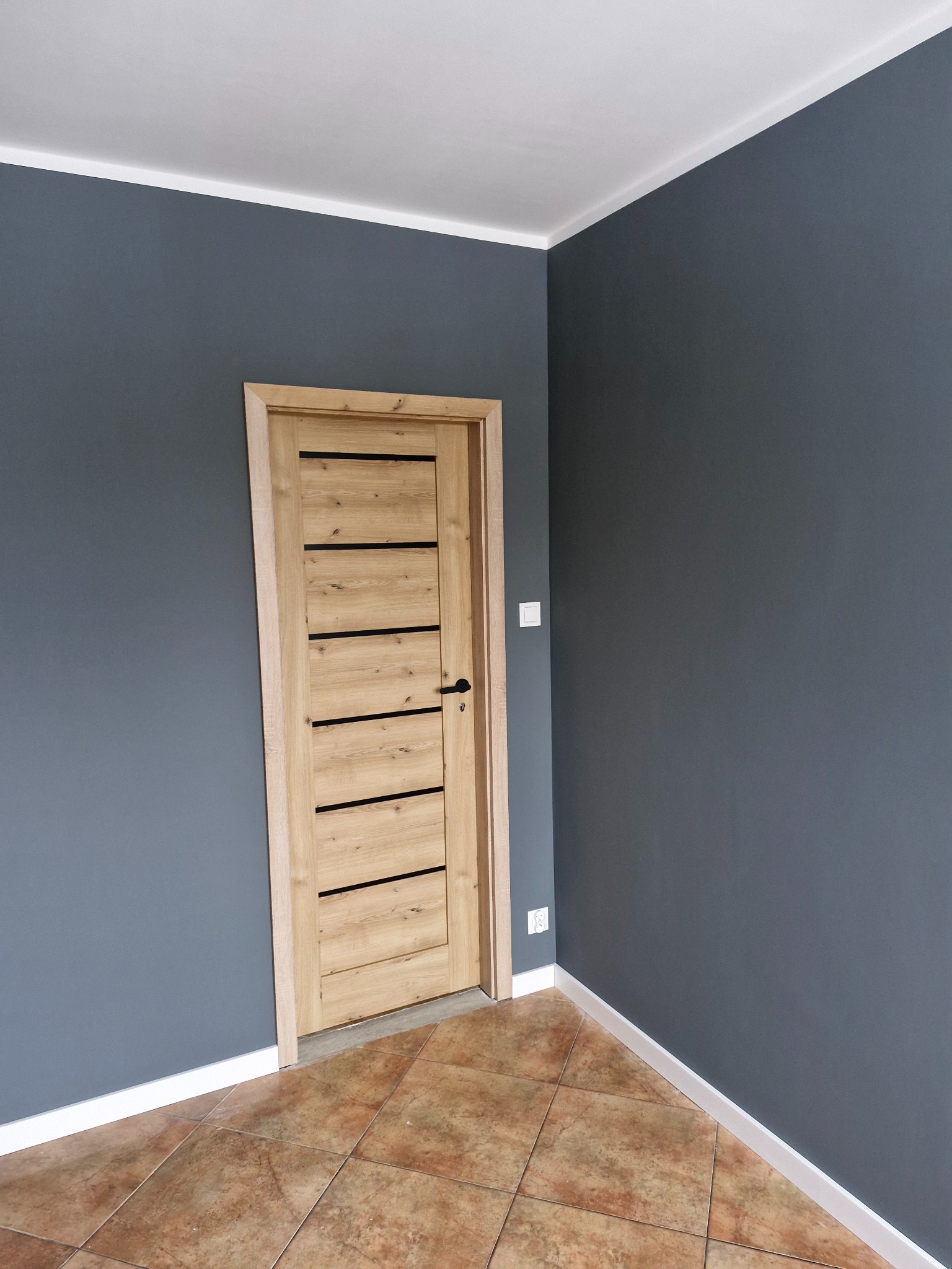 Wnętrze po remoncie: drewniane drzwi z czarnymi akcentami na tle grafitowej ściany, brązowa podłoga z płytek, białe listwy przypodłogowe i sufitowe.