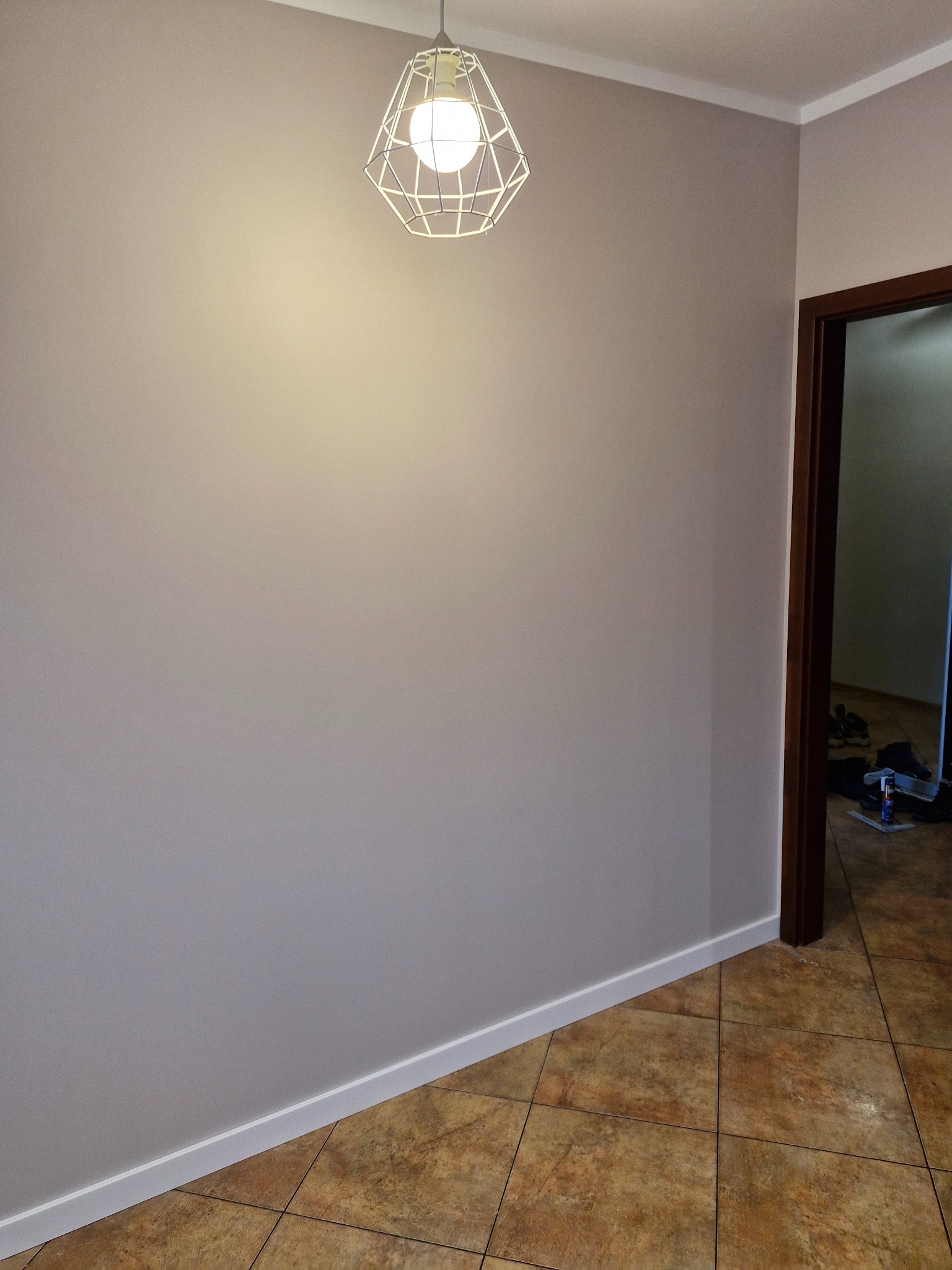 Pomalowana na szaro ściana z białą listwą przypodłogową i ozdobną lampą. W tle widoczne otwarte drzwi z brązową futryną i fragment podłogi z płytek.