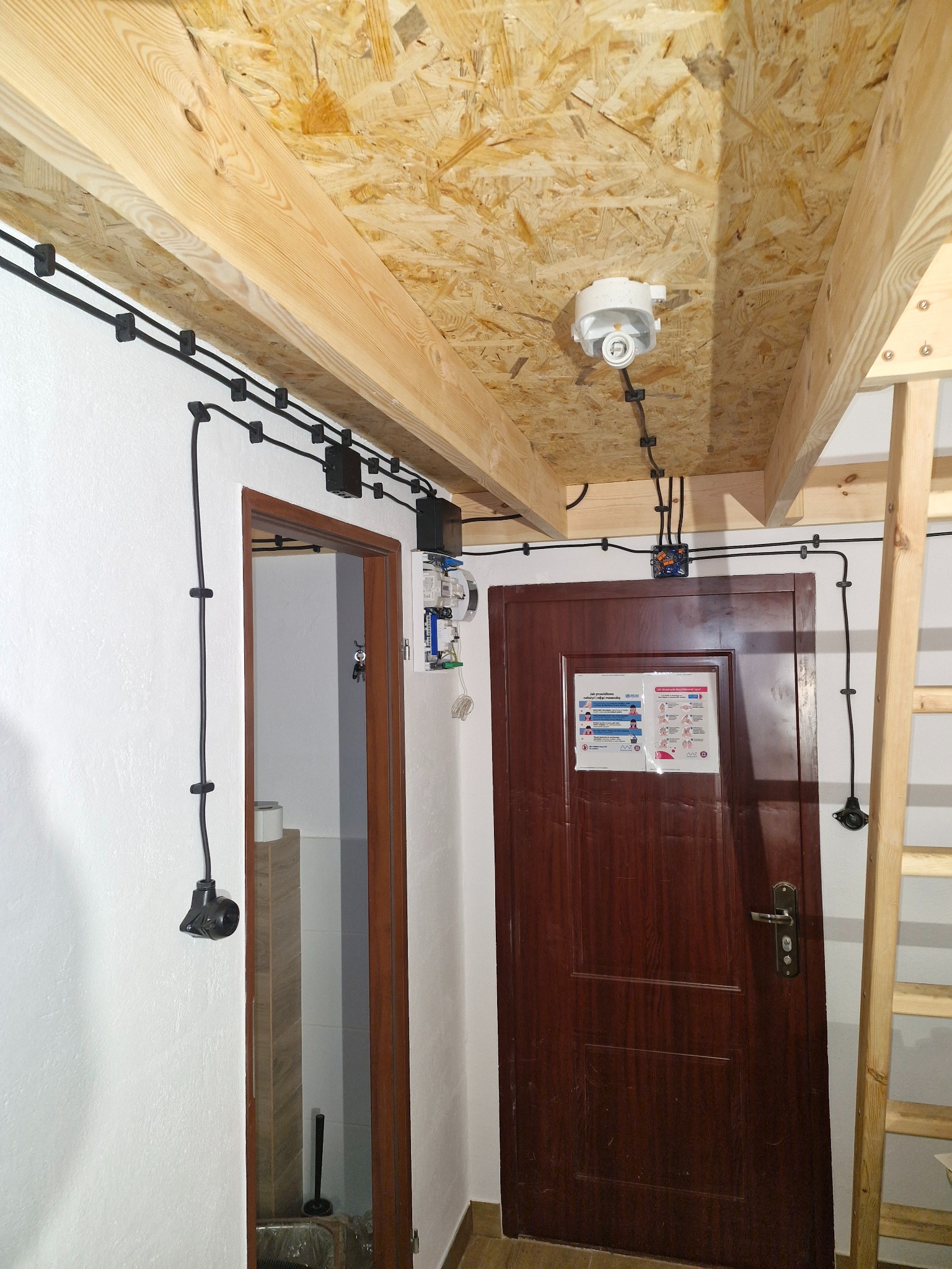 Instalacja elektryczna natynkowa w pomieszczeniu z drewnianym stropem, widoczne kable, puszki, licznik i oprawa oświetleniowa. Drzwi i drabina w tle.