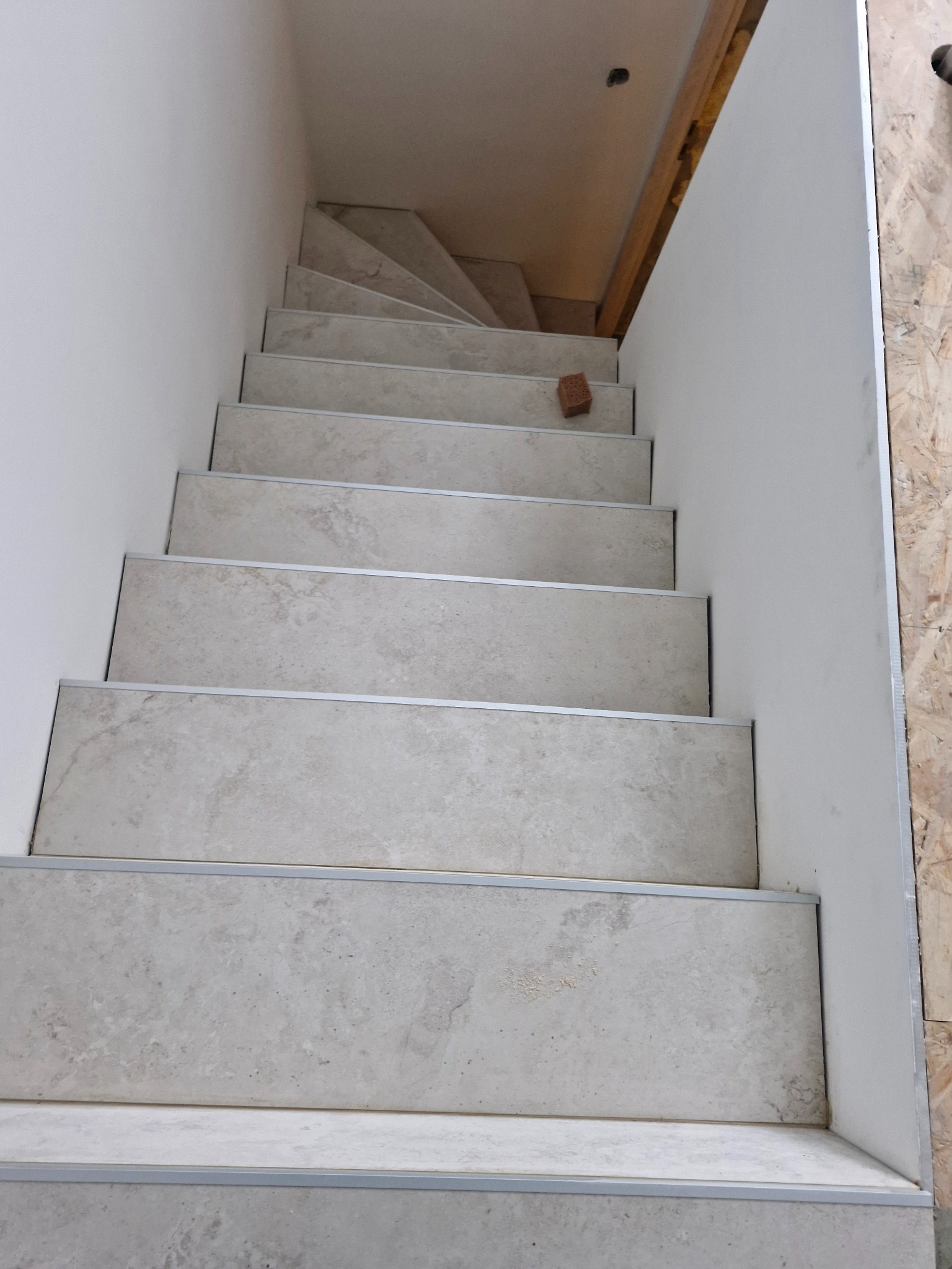 Wykończone schody wewnętrzne z jasnymi, kamiennymi płytkami i srebrnymi listwami krawędziowymi, widok z góry. Na jednym ze stopni leży cegła.