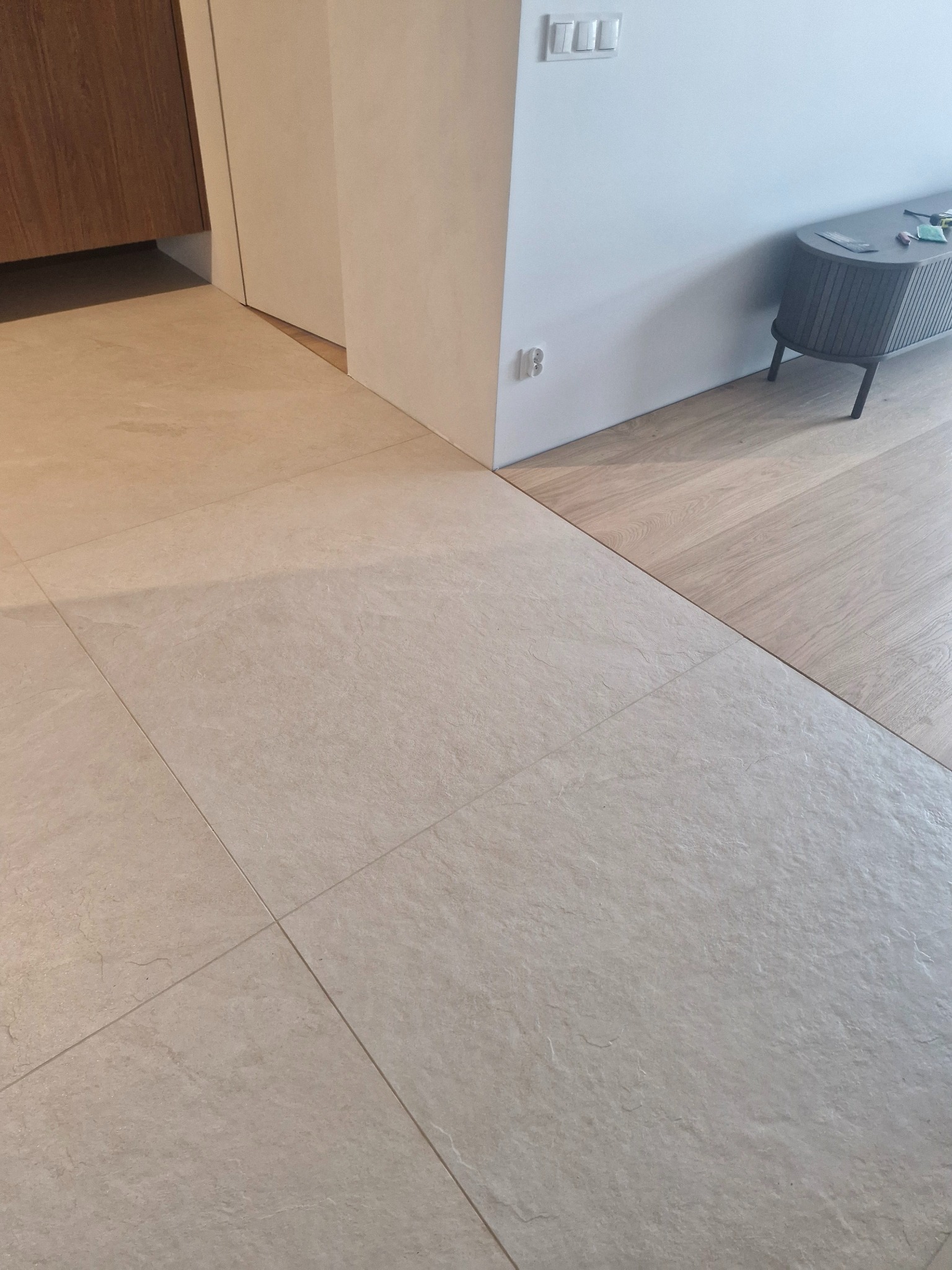 Beżowe, matowe płytki podłogowe ułożone w kwadraty, kontrastujące z drewnianą podłogą w tle. Widoczny fragment szafki i ściana z włącznikami.