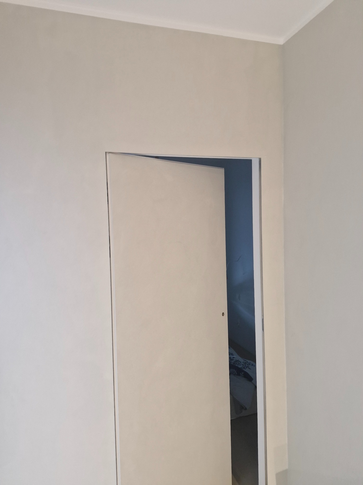 Otwarta, beżowa, minimalistyczna ściana z ukrytymi drzwiami w pokoju. Widoczny fragment wnętrza z pościelą w tle. Świeżo pomalowane ściany w neutralnych barwach.