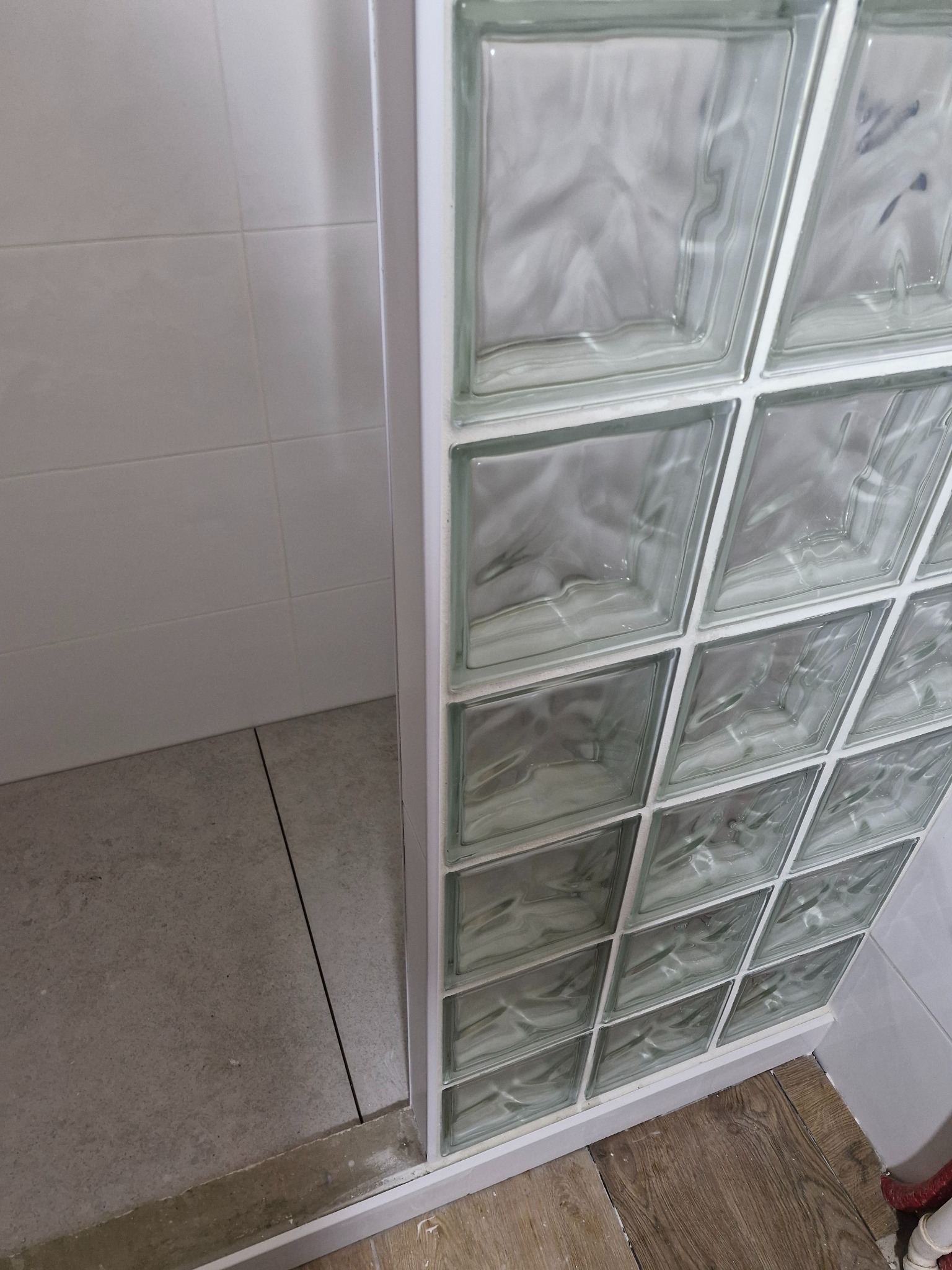 Szklana ścianka działowa z luksferów w łazience, widok z bliska na połączenie z podłogą i ścianą. Minimalistyczny design, jasne płytki.