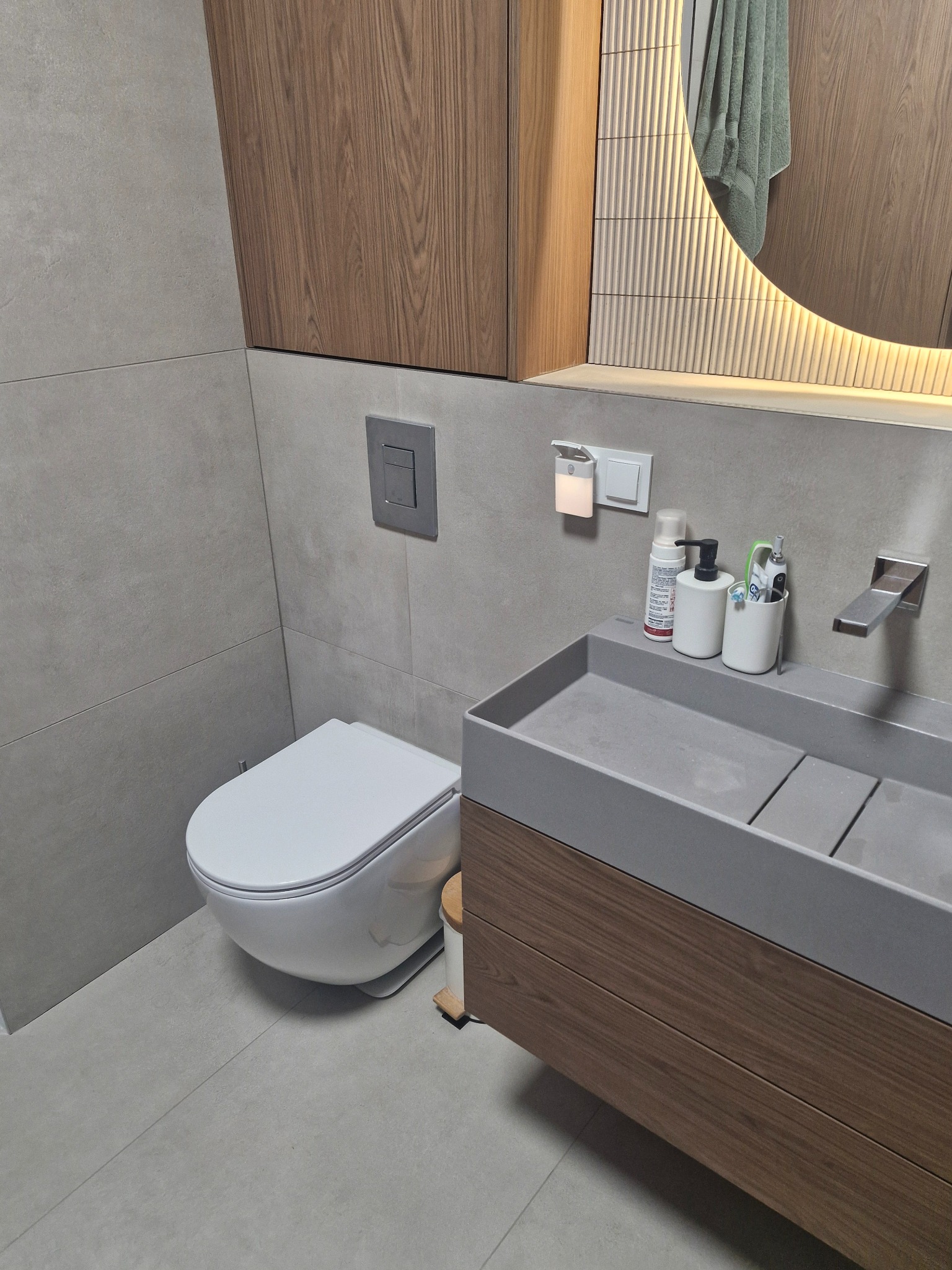 Nowoczesna łazienka z podwieszaną toaletą, szarą umywalką i okrągłym lustrem z podświetleniem LED. Drewniane szafki wiszące i szare płytki tworzą spójną całość.
