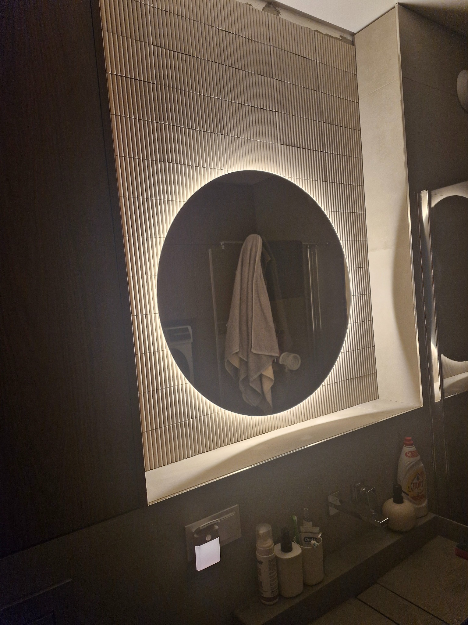 Wykończona łazienka w Toruniu: ściana z dekoracyjną glazurą, okrągłe lustro z podświetleniem LED, nowoczesna umywalka z armaturą i akcesoriami.