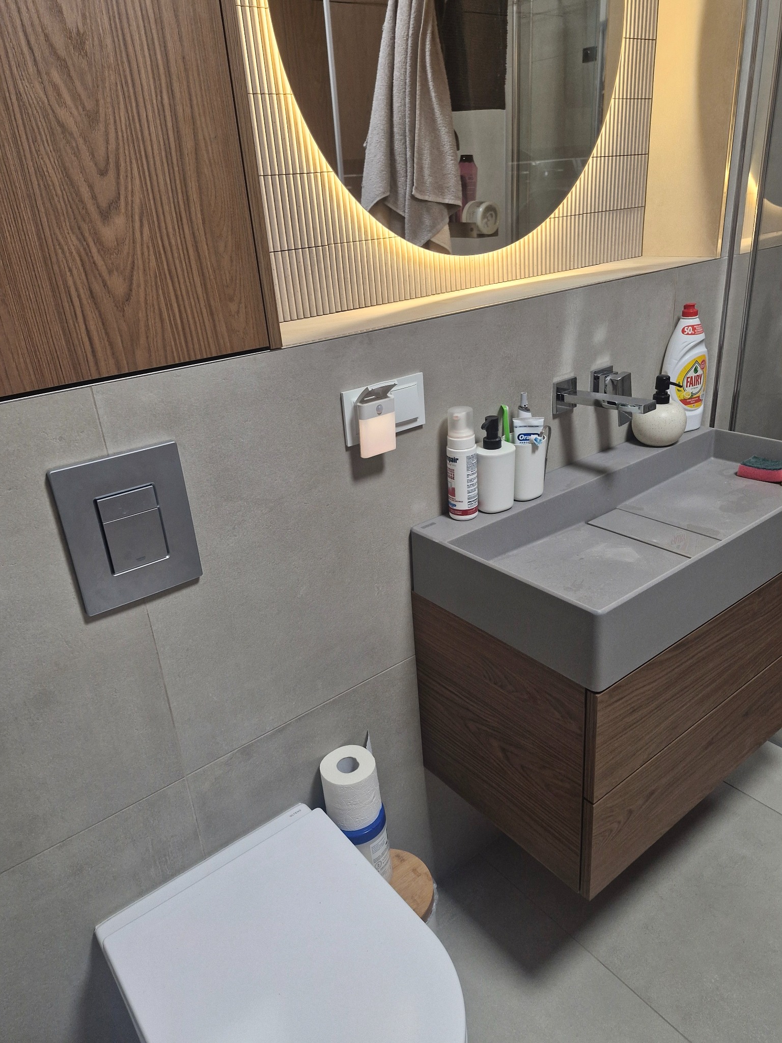 Nowoczesna łazienka z podświetlanym lustrem, umywalką z szafką z drewnianym fornirem i wiszącą toaletą. Szare płytki na ścianach i podłodze.